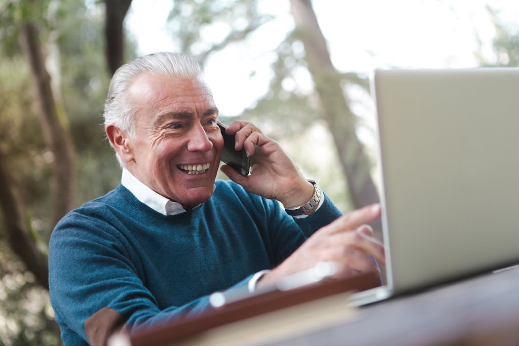 Cheerful senior man working remotely, talking on phone and using laptop outdoors.